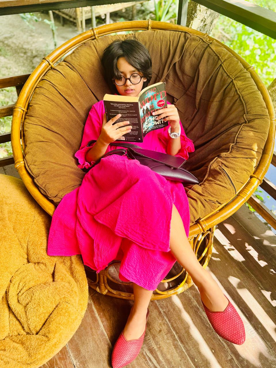 Drisana reading a book in a comfortable rattan chair, wearing a bright pink dress